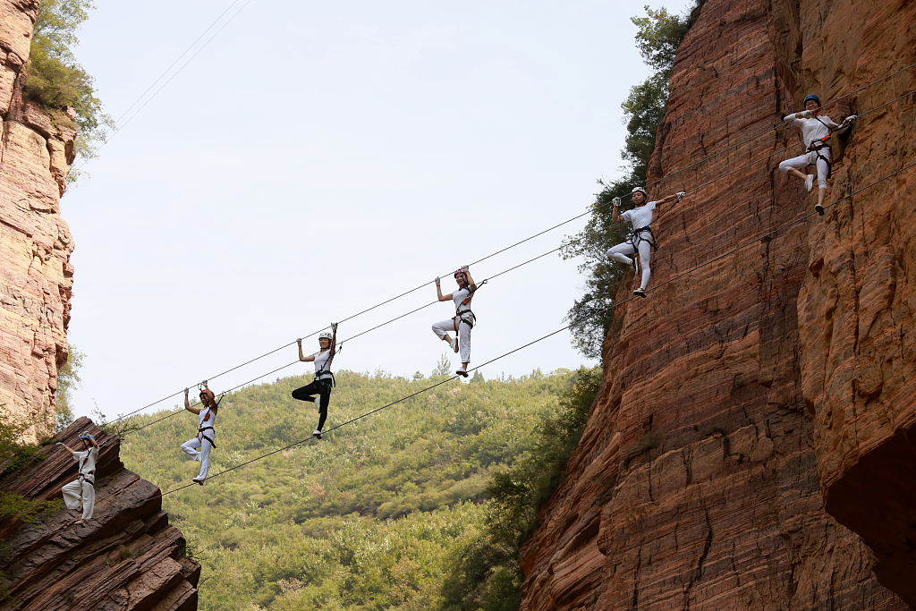 Berdiri di tepi tebing terjal dan mengenakan pakaian yoga yang nyaman mereka dengan percaya diri menyeimbangkan diri di atas kabel baja yang tergantung di permukaan tebing